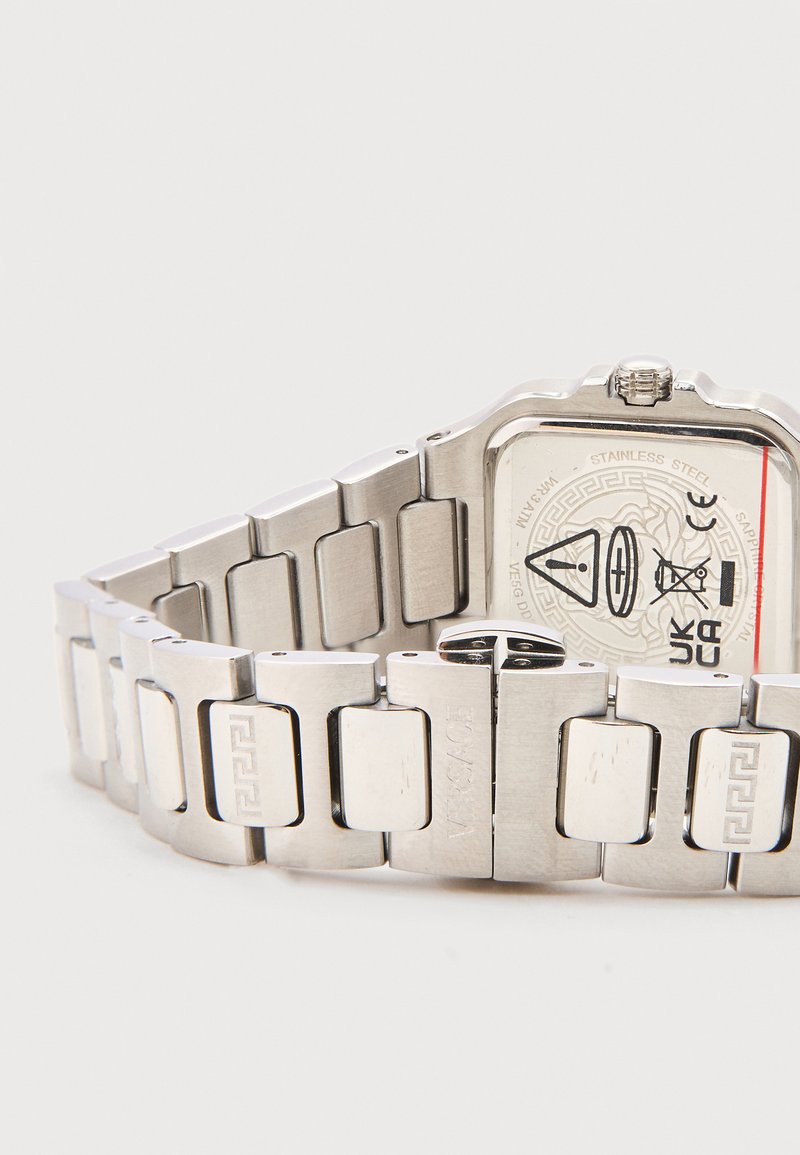 Silver metal watch band with engraved rectangular links and a square watch face showing safety symbols and certification marks.