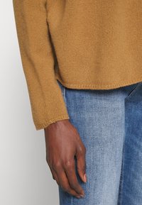 Brązowy sweter z dzianiny o strukturalnej powierzchni, charakteryzujący się luźnym krojem i prążkowanymi mankietami, w połączeniu z jasnoniebieskimi dżinsami.