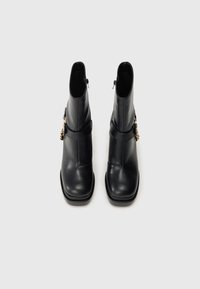 Fekete bőr bokacsizma pár, négyzet alakú orral és aranylánc részletekkel a külső oldalán, fehér háttéren, felülnézetből bemutatva.