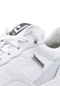 Zapatilla deportiva de cuero blanco con cordones blancos, lengüeta perforada, placa metálica con el logotipo y etiqueta de tela con el logotipo.
