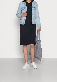 Jeansjacka, svart huvad klänning, vita sneakers och en randig tote-väska. Funktioner inkluderar kontrasterande färger och varierande texturer.