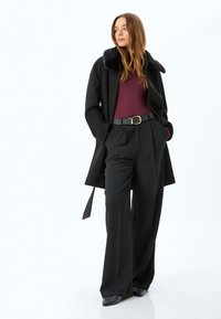 Cappotto nero con colletto in pelliccia sintetica, abbinato a un top bordeaux e pantaloni neri a gamba larga. Presenta una vita con cintura e un design fino al ginocchio.