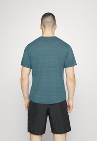 Man with short hair wearing a teal short-sleeve shirt and black shorts, standing facing away against a plain light background.