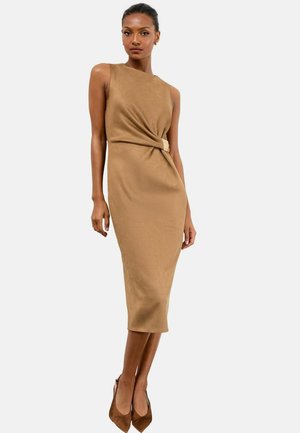 Vrouw in een mouwloze tan midi-jurk met zijdelings gespdetail en brown puntige hakken, staande tegen een plain witte achtergrond.
