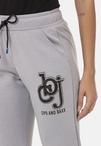 Helle graue Sweatpants mit einem schwarzen Drucklogo, seitlichen Taschen und einem Kordelzug mit bunten Spitzen. Weicher Stoff.