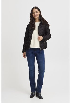 Zwarte donsjas met chevronpatroon en ritssluiting, gecombineerd met een licht beige trui en blauwe spijkerbroek, gedragen met zwarte enkellaarsjes.