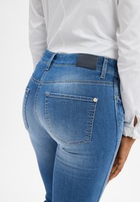 Blue denim jeans feature a slim fit with a mid-rise waist, back pockets, and a leather patch on the waistband. The fabric has a slight fade effect.