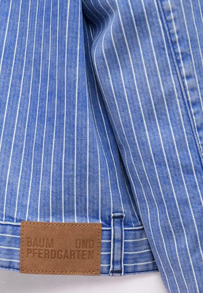 Ύφασμα με μπλε και λευκές ρίγες και λεία υφή· μια καφέ δερμάτινη ετικέτα φέρει ανάγλυφο κείμενο «BAUM UND PFERDGARTEN».
