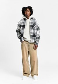 Karo-Flanellhemd in Schwarz, Weiß und Grau; grauer Hoodie darunter; beigefarbene weite Hose; weiße Sneaker; lockere Passform.