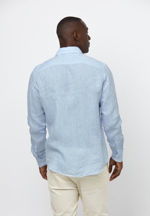 Mand set bagfra iført en lyseblå langærmet skjorte og beige bukser mod en ensartet hvid baggrund.