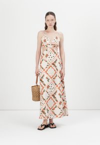 Maxi-jurk met een bloemenprint in oranje en groen op een crèmekleurige achtergrond. Voorzien van een V-hals, uitsnijdingen aan de zijkanten en dunne schouderbandjes. Geweven mand tas en zwarte sandalen.