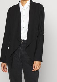 Veste légère noire présentant un devant drapé, des manches longues et une fermeture à crochet et boucle. Portée par-dessus une chemise blanche à col.