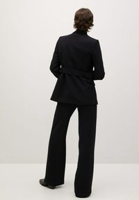 Traje negro a medida con cintura ceñida, hombros estructurados y pantalones de pierna ancha, con una textura suave y costuras detalladas.
