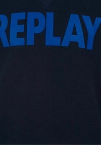 Lettere blu in grassetto maiuscole che formano la parola "REPLAY" su uno sfondo di tessuto scuro, probabilmente un primo piano di una maglietta o di un felpa.