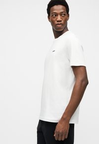 Mladý muž v jednoduchém bílém tričku s malým černým nápisem na hrudi, dívající se přes levé rameno na jednoduchém pozadí.