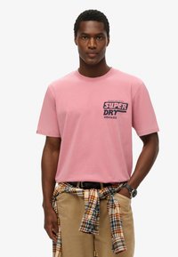 Camiseta de algodón rosa claro con mangas cortas, que presenta un logo en azul oscuro de "SUPER DRY" en el lado izquierdo del pecho. Llevada con pantalones beis y una camisa de cuadros atada a la cintura.