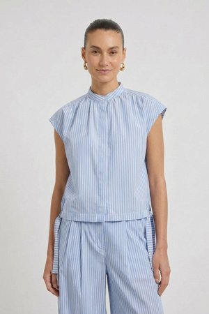 Femme aux cheveux tirés en arrière portant des boucles d’oreilles créoles en or, un chemisier à manches courtes bleu clair et blanc à rayures verticales et un pantalon assorti.