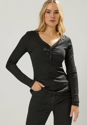 Blondynka w dopasowanym czarnym prążkowanym długim rękawem z guzikami, pozująca z jedną ręką w kieszeni na jednolitym tle.