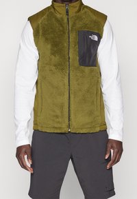 Olivgrön väst med en fluffig textur, svart dragkedjeficka och kontrasterande slät tyg vid axlarna. Buren över en vit långärmad skjorta.