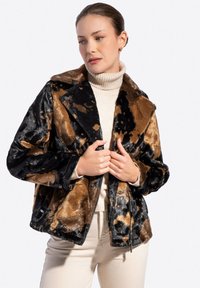 Kobieta poprawiająca czarną i brązową wzorzystą kurtkę z futerka na beżowy sweter z golfem i jasne spodnie.