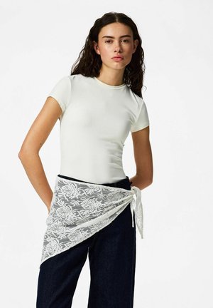 Young woman with dark curly hair wears a fitted white short-sleeve shirt, dark jeans, and a white floral lace wrap tied at the waist.