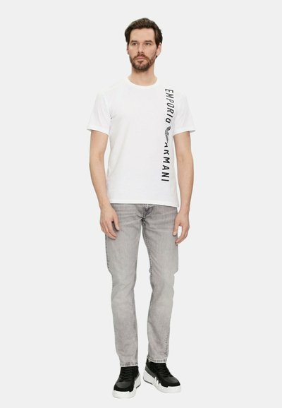 T-shirt bianco in cotone con stampa verticale nera del logo. Abbinato a jeans grigi chiari e sneakers nere, completando un look casual.