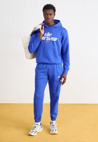 Felpa Nike blu con tasca frontale e stampa del logo, abbinata a pantaloni joggers blu coordinati. Il modello tiene una borsa a tracolla beige e indossa sneakers con motivo.