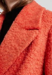 Femme portant un manteau texturé corail orange avec un large revers sur un haut noir, gros plan sur le col et la zone de l'épaule.