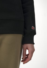 Sudadera negra con puño acanalado, con el logo de Lacoste en rojo y blanco. Combínala con pantalones texturizados color beige, mostrando una mano descansando ligeramente.