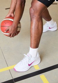 Bílé sportovní boty s fialovým logem Nike, doplněné bílými ponožkami. Basketbal je držen oběma rukama, umístěn na podlaze hřiště.