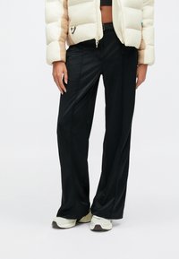 Oseba, oblečena v črne široke hlače, bele superge in bež-bel pufasto jakno, stoji ob enobarvnem ozadju.