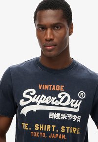 Navy blauwe katoen t-shirt met een ronde hals, met witte en oranje "SuperDry" tekst en extra grafische elementen in een versleten stijl.