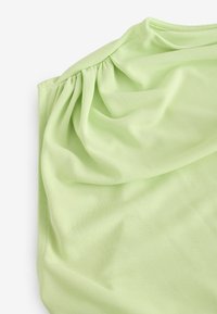 Top suave de color verde claro con tejido drapeado y detalles fruncidos en el hombro. La textura suave y elástica aumentan la comodidad.