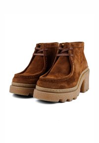 Braune Wildleder-Ankelstiefel mit einer dicken, strukturierten Sohle. Merkmale sind eine runde Zehenpartie, zwei braune Schnürsenkel und ein klobiger Absatz für Stabilität.