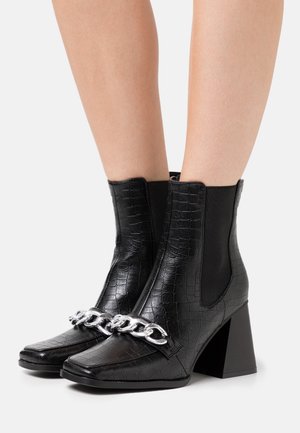 Mustat nilkkurit krokotiilipintaisesta nahasta, joissa on neliönmuotoinen kärki, metalliketjuverhoilu ja paksu lohkopohja.