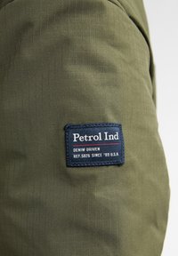 Grüner Stoff mit einem Karomuster, der ein blaues Patch mit der Aufschrift "Petrol Ind" zeigt, mit roten und weißen Schriftzügen, die Markennamen und Referenzinformationen detailieren.