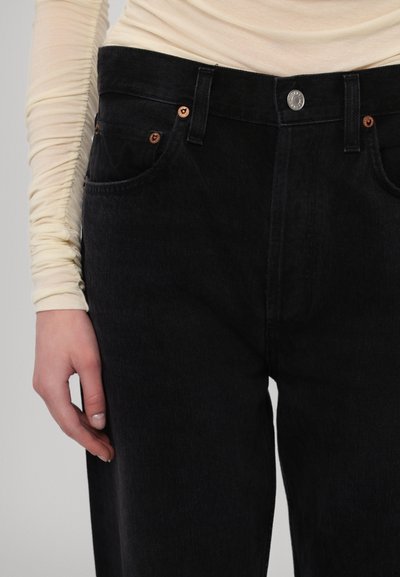 Black denim jeans with a high waist, featuring copper button accents, front pockets, and a smooth texture. Styled with a fitted cream long sleeve top.