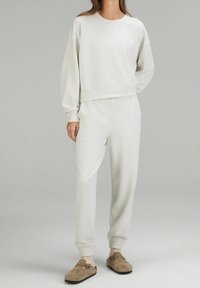 Mujer vestida con un suéter de manga larga color beige claro y pantalones jogger a juego, con calcetines beige y zapatos de lona marrones, de pie sobre un fondo gris.