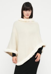 Donna che indossa un poncho color crema con bordi di pelliccia bianca sopra un vestito nero a maniche lunghe, in piedi davanti a uno sfondo bianco semplice.