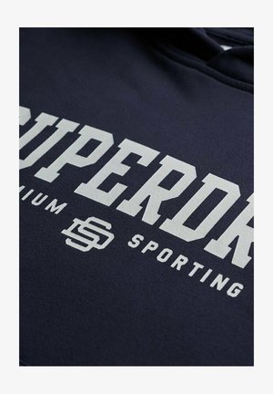 Marineblauwe hoodie met grote witte letters met de tekst "SUPERDRY" en kleinere tekst "SPORTING." Zachte stof met een gestructureerde afwerking.