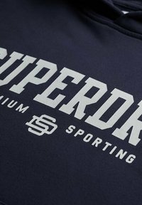 Marineblauwe hoodie met een groot wit "SUPERDRY" logo, met daaronder kleinere teksten "premium" en "sporting", gemaakt van zachte stof.