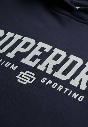 Marineblauwe hoodie met een groot wit "SUPERDRY" logo, met daaronder kleinere teksten "premium" en "sporting", gemaakt van zachte stof.