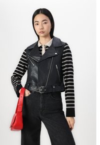 Black leather biker vest with a belted waist, featuring zippers and lapels. Worn over a black and white striped long-sleeve top, paired with black jeans.