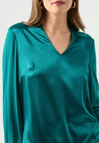 Blusa a maniche lunghe in satin verde acqua con scollatura a V e vestibilità morbida. Tessuto liscio con una leggera lucentezza, dotata di una tasca laterale.