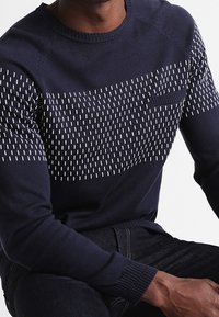 Donkerblauw gebreid sweater met een gestructureerd bovenstuk met witte verhoogde stikpatronen en een borstzak. Geribbelde mouwen en zoom.