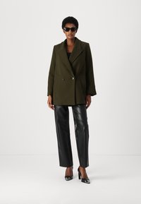 Palton de culoare verde măslin, cu două rânduri de nasturi și rever mari, combinat cu pantaloni din piele neagră și pantofi cu toc înalt.