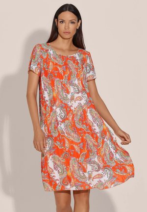 Vrouw draagt een oranje-witte paisley jurk tot op de knieën met korte mouwen, staand tegen een effen beige achtergrond.
