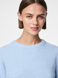 Světle modrý pletený svetr s žebrovaným vzorem a kulatým výstřihem. Modelka má rovný bob a minimalistické náušnice, které zdůrazňují texturu oděvu.