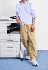 Ljusblå hoodie med vita detaljer, beige löst sittande byxor och vita träningsskor, stående bredvid en skrivare.