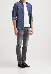 Ciemnoniebieska koszula z długim rękawem zapinana na guziki noszona na białej koszulce, w zestawieniu z szarymi dżinsami i czarnymi sneakersami. Prosty wzór z kieszeniami na koszuli.